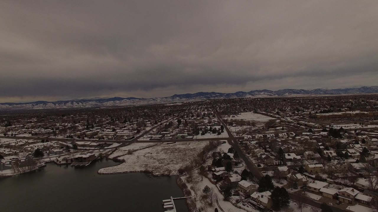 Aerial view in Jefferson County, Colorado showing a dark lake, snowy shoreline, residential streets, distant mountains, and no visible people at the start of the clip.