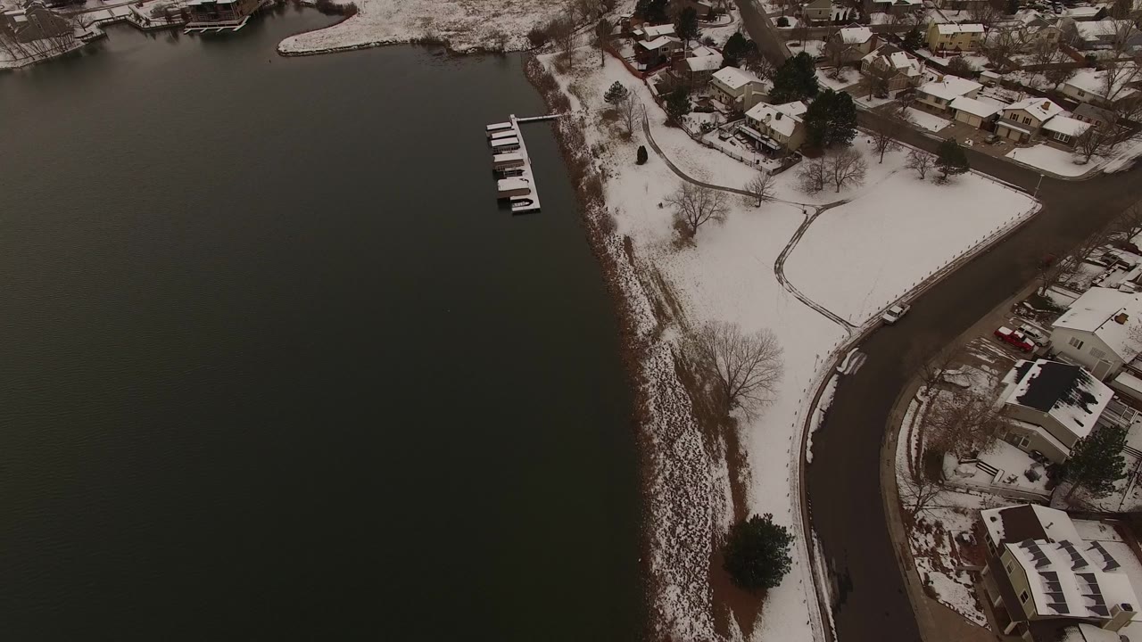 High aerial view in Jefferson County, Colorado with a curved road, snow-dusted houses, bare trees, shoreline rocks, and dark water filling the right side, with no visible people.