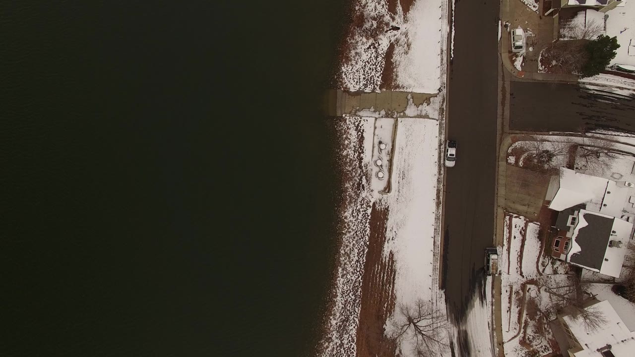 Top-down aerial view of dark water, snowy shoreline, road, and lakeside houses.
