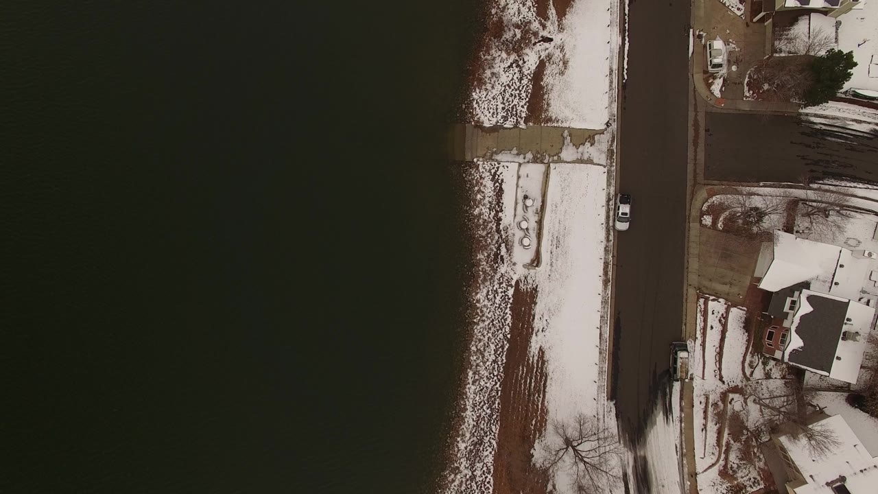 Overhead Jefferson, Colorado view with dark water filling the left side, a snow-dusted bank beside a road, and private homes and yards on the right, with no visible people.