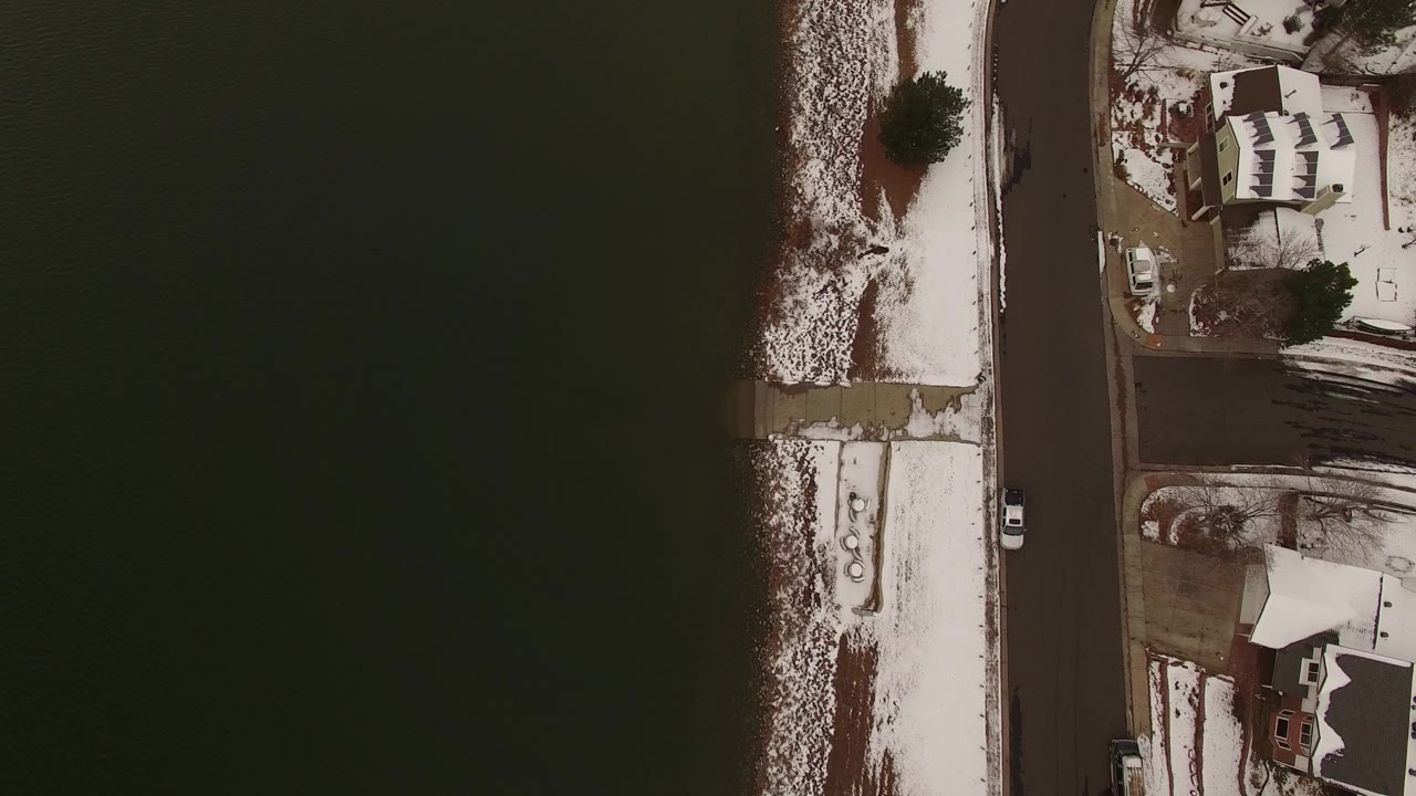 Overhead aerial frame in Jefferson, Colorado showing dark water, a snow-lined shore, a straight residential road, rooftops, bare trees, and no visible people.
