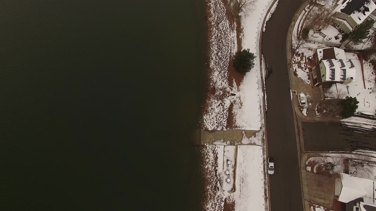 Top-down snowy shoreline with houses, road, and dark water.