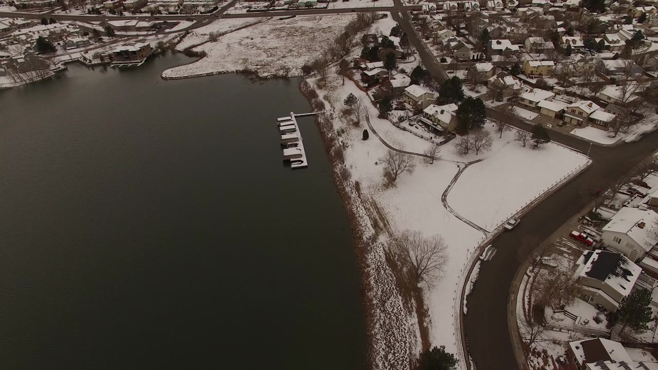 Aerial view over a dark lake beside snow-covered paths, docks, bare trees, streets, and private homes in Jefferson, Colorado, with no clearly visible people.