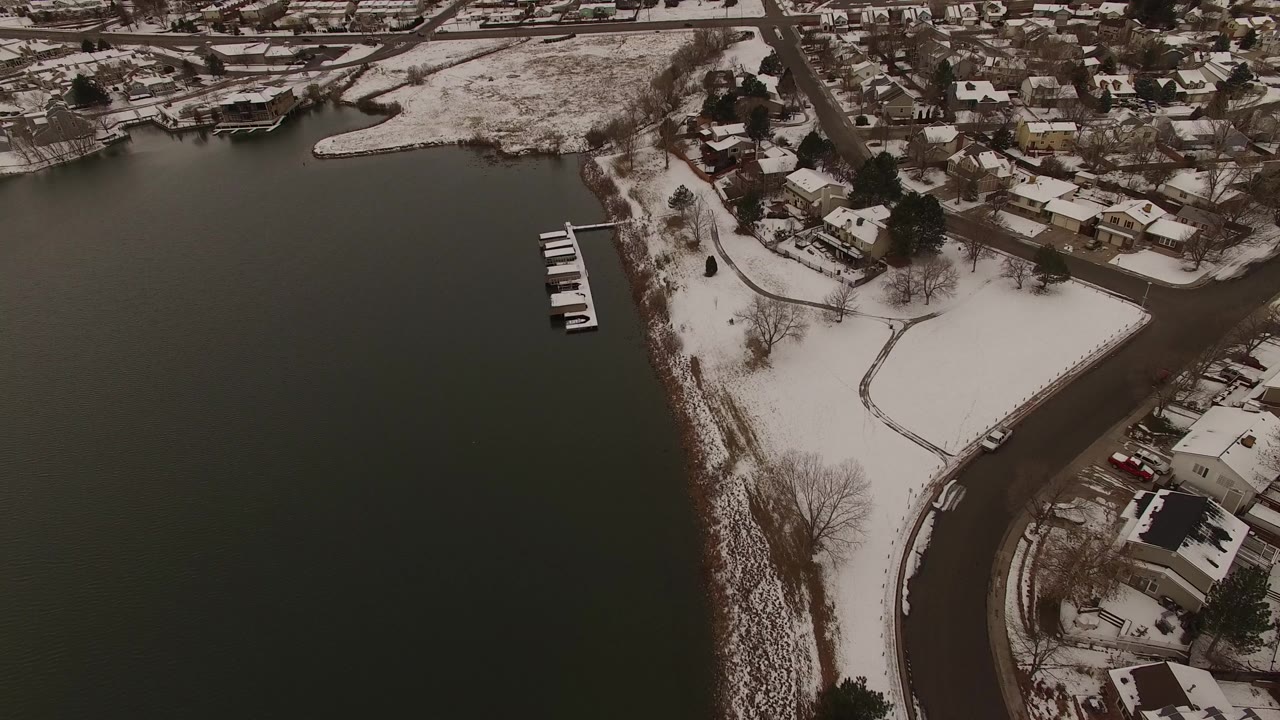 A high view of the Jefferson, Colorado lakefront shows dark water, a white path through snow, small docks, bare trees, nearby roads, and private homes with no clearly visible people.