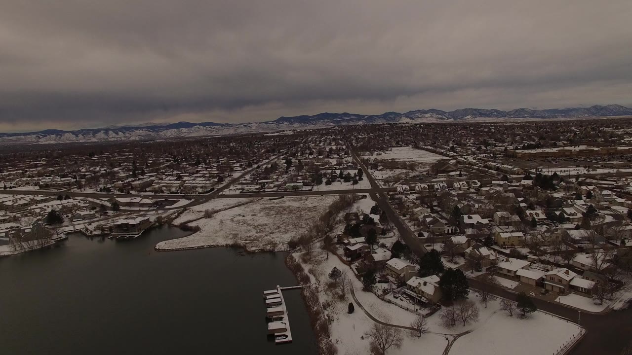 Opening aerial frame over Jefferson County, Colorado, showing dark lake water, snow-covered shore, houses, roads, distant mountains, and no visible people.