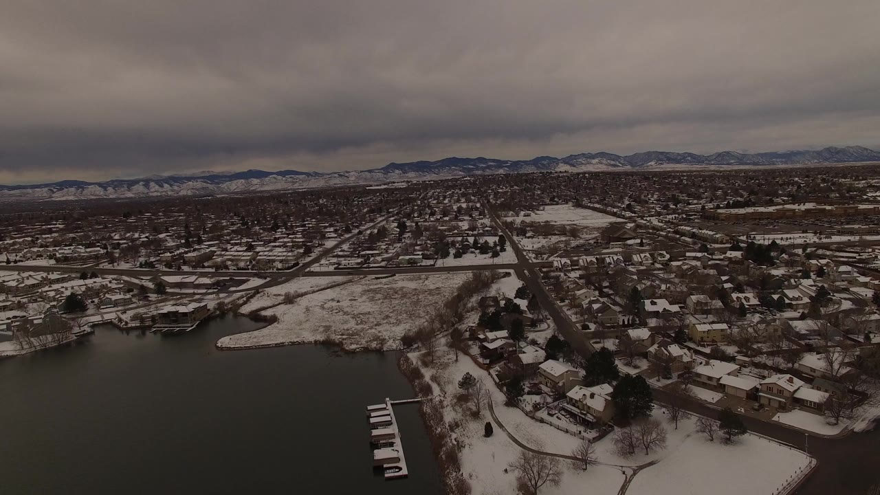 Aerial frame in Jefferson County, Colorado, with the lakeshore lower in frame, clustered homes and streets in the center, distant mountain ridges, and no visible people.