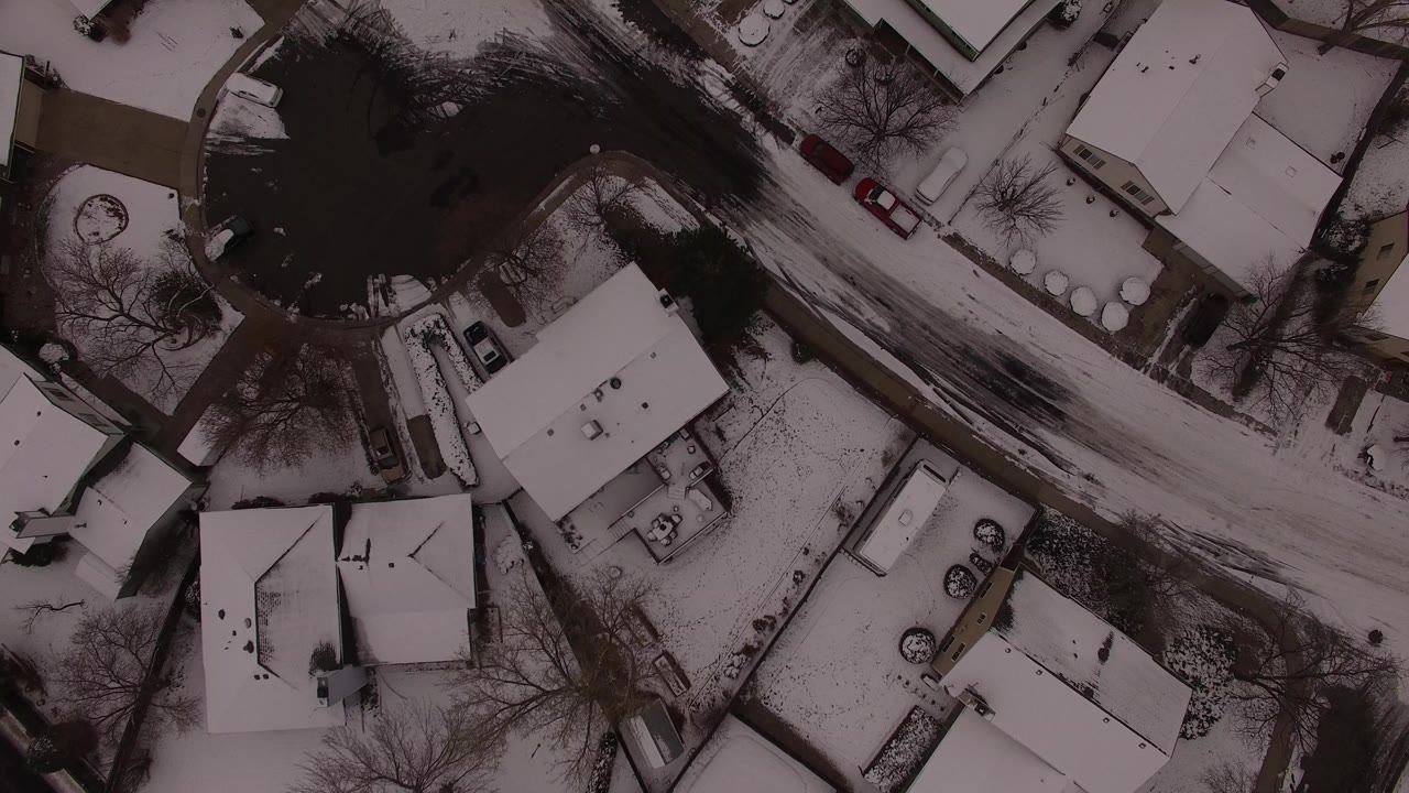 Overhead frame from Jefferson County, Colorado showing white roofs, dark tree cover, a snow-lined curved road, parked vehicles, and no visible people.