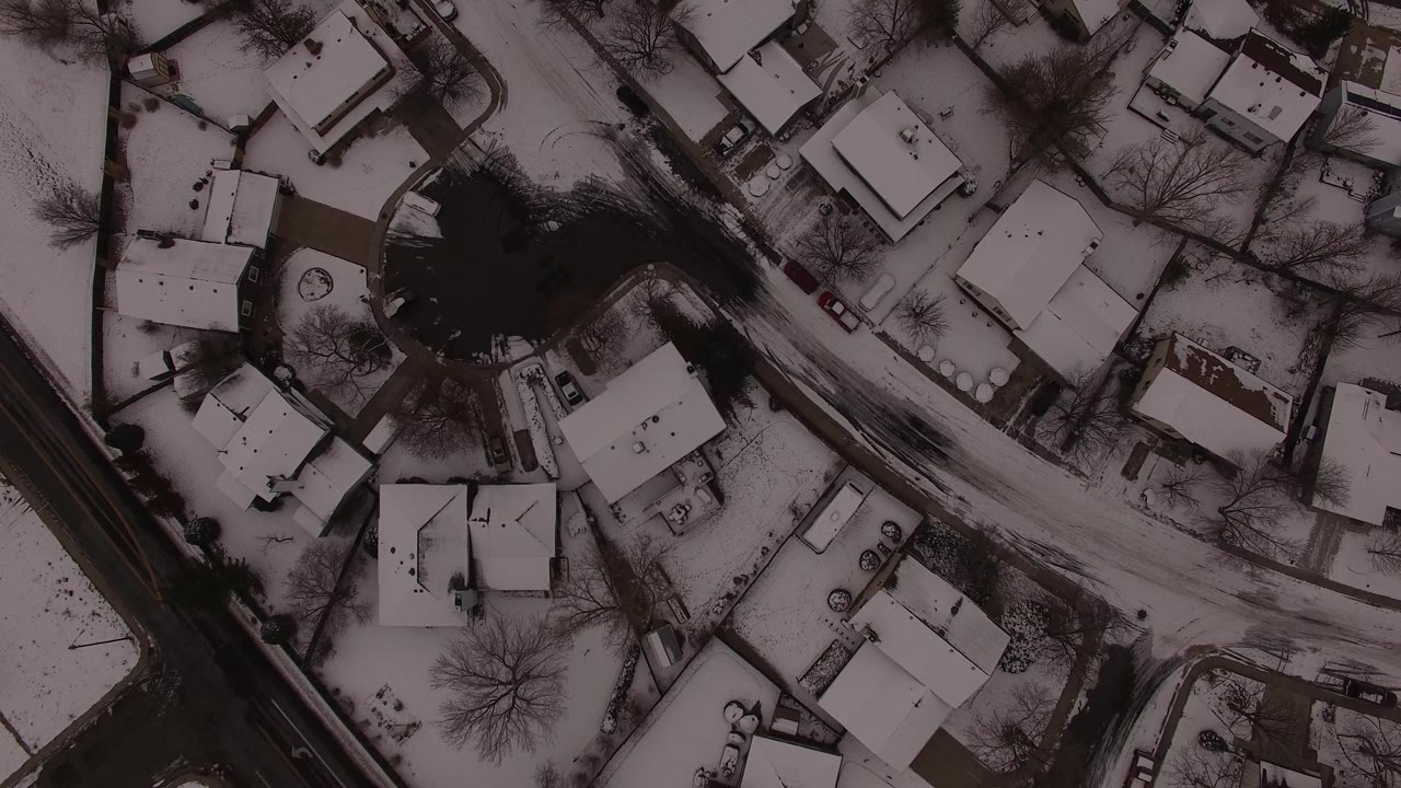 Overhead view in Jefferson County, Colorado, showing snow-covered homes, driveways, a diagonal road, bare trees, and no visible people at the opening moment.