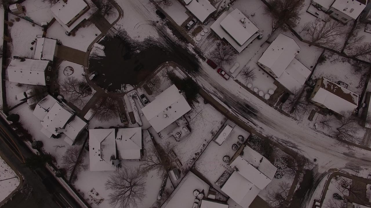 Overhead aerial frame of a snow-covered residential area in Jefferson County, Colorado, with a diagonal road, white rooftops, bare trees, and no visible people.