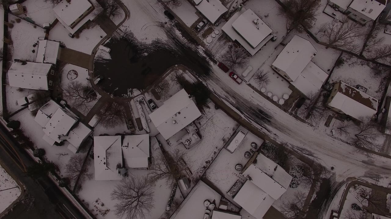 Straight-down aerial view over Jefferson County homes where the dark pond or tree mass sits near the upper left and the snowy road crosses the frame, with no visible people.