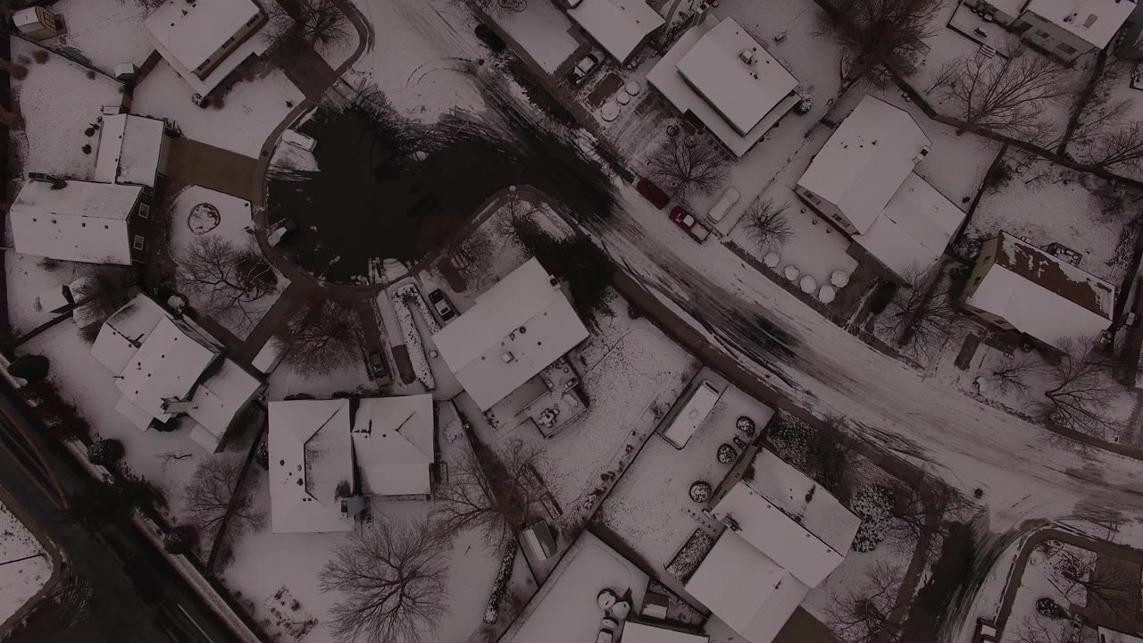 Aerial snowy subdivision with rooftops and a curving road.