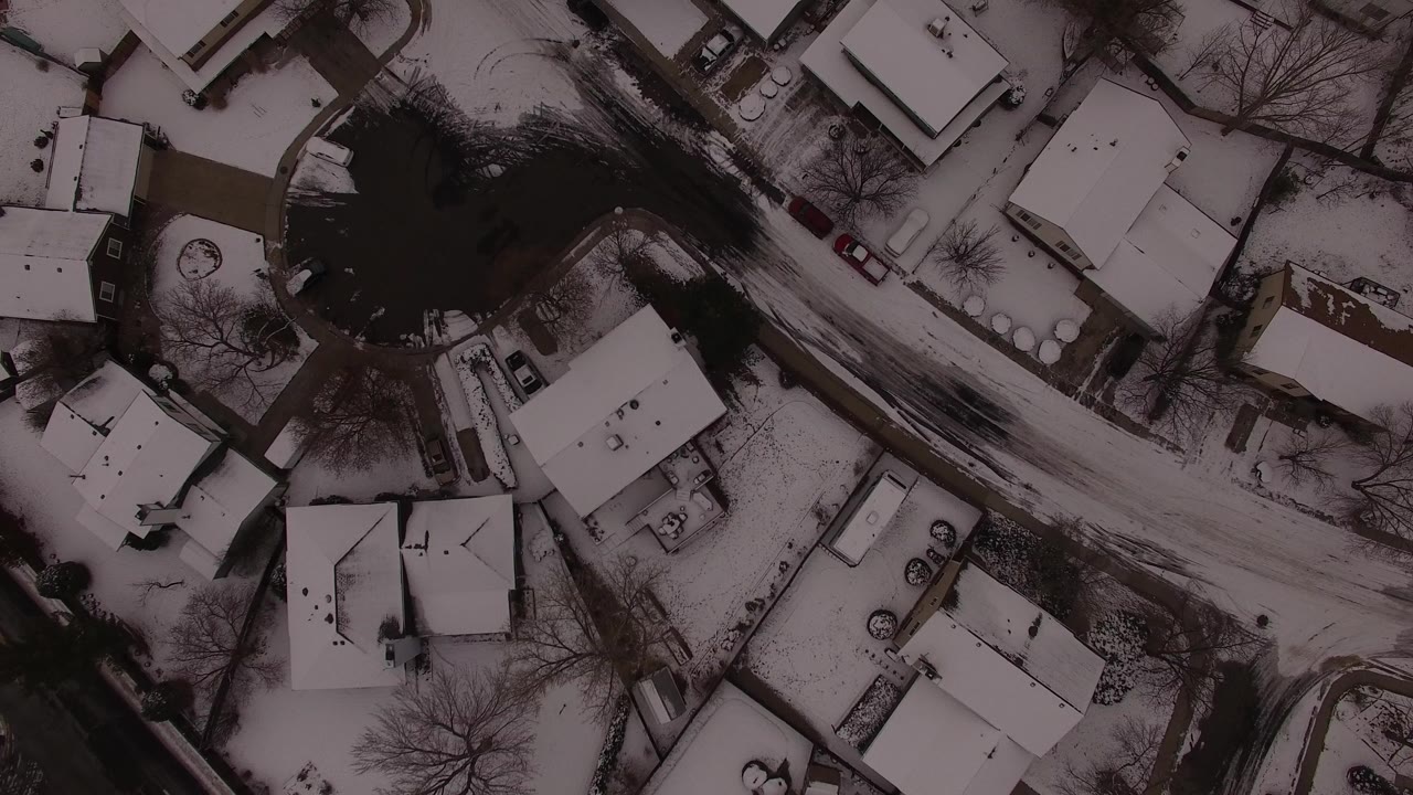 Top-down aerial frame over Jefferson County, Colorado showing snow-covered homes, bare trees, driveways, parked cars, and a dark curving street with no visible people.