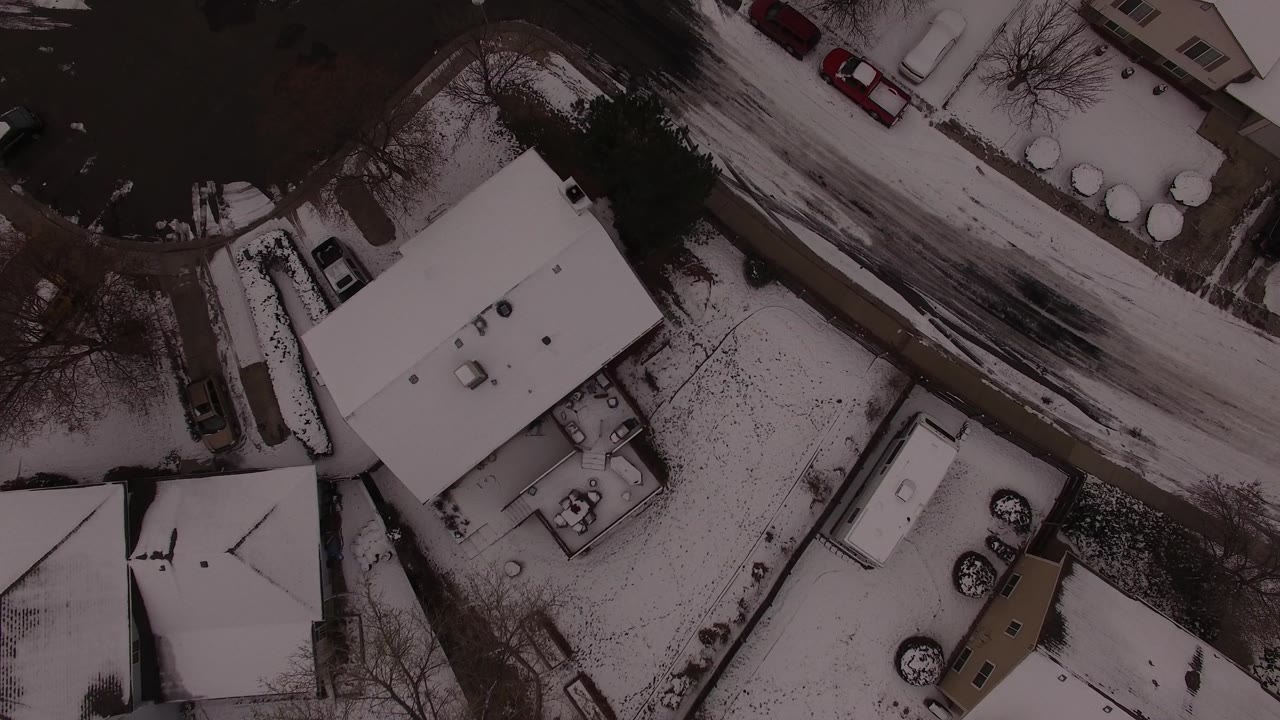 Overhead Jefferson County, Colorado frame with a large snow-covered roof near center, a plowed residential road above it, parked cars, yard fencing, and no visible people.