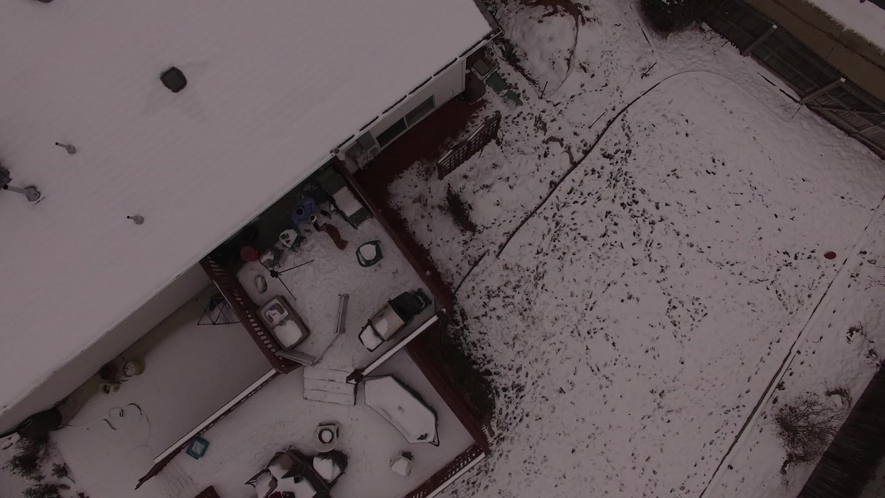 Overhead frame in Jefferson County, Colorado showing snow-covered house roofs, a fenced patio area, parked vehicles, and a white yard with no visible people.