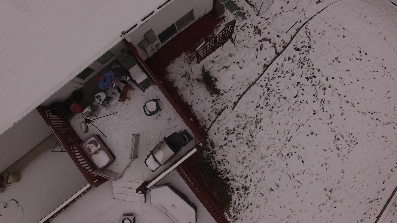 Aerial view in Jefferson County, Colorado looking down on the house corner, red deck, patio objects, scattered chairs, and snow-covered yard with no visible people.