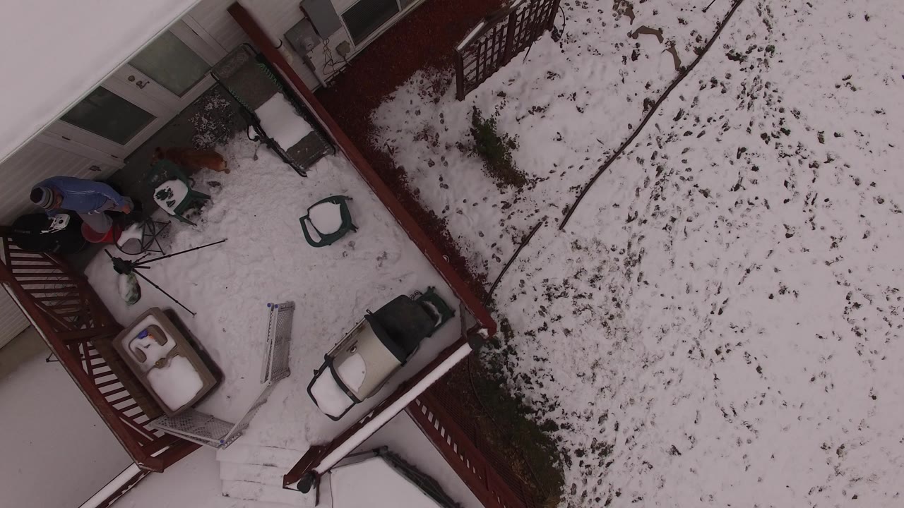 Overhead snowy yard beside a red deck with a man and dog near the house.