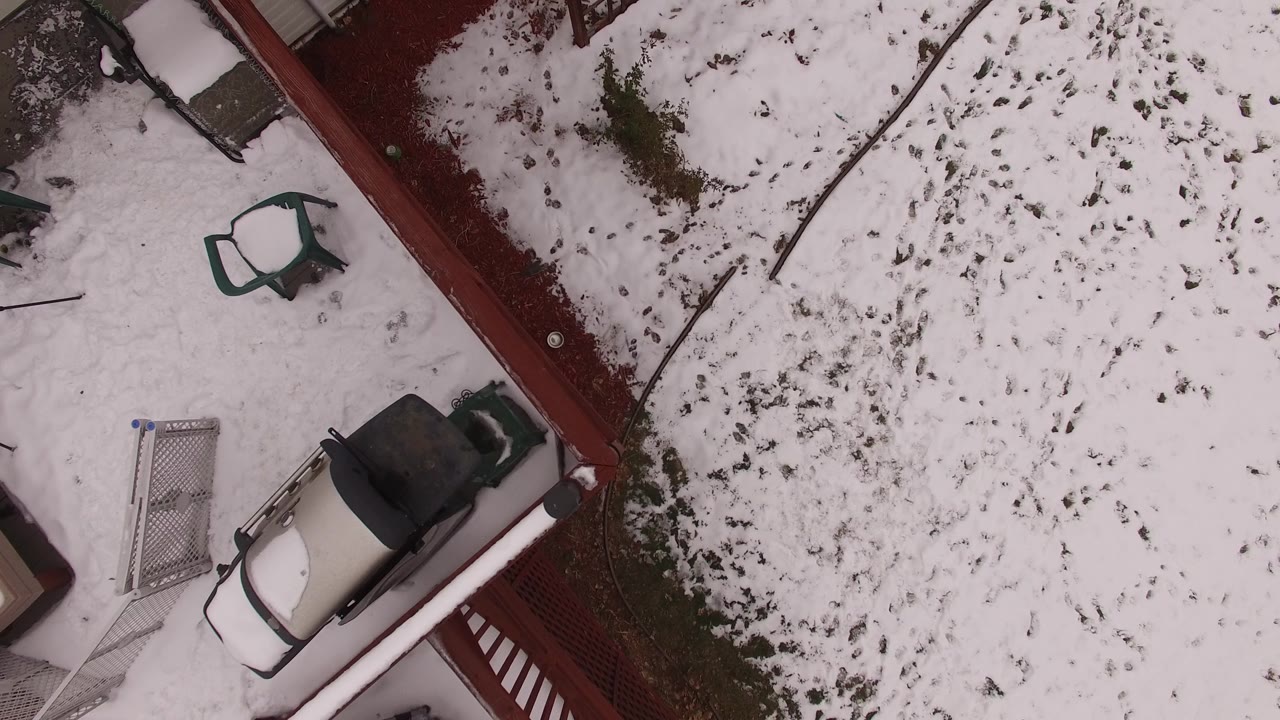 Overhead frame of the Jefferson County, Colorado yard showing the red fence line, green patio chair, pale vehicle, metal gate, patchy snow, and no visible people.