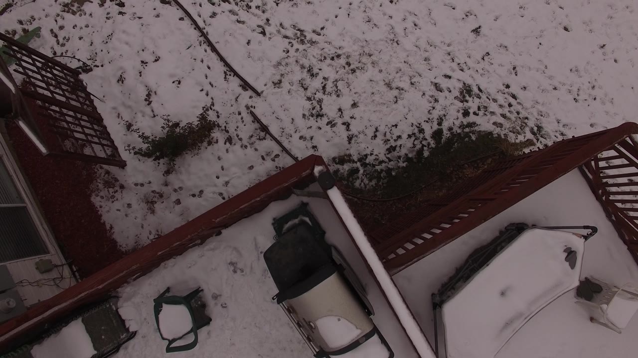 Aerial frame over a Jefferson County, Colorado backyard showing snow, exposed ground, a red fence or deck rail, part of a house wall, and no visible people.
