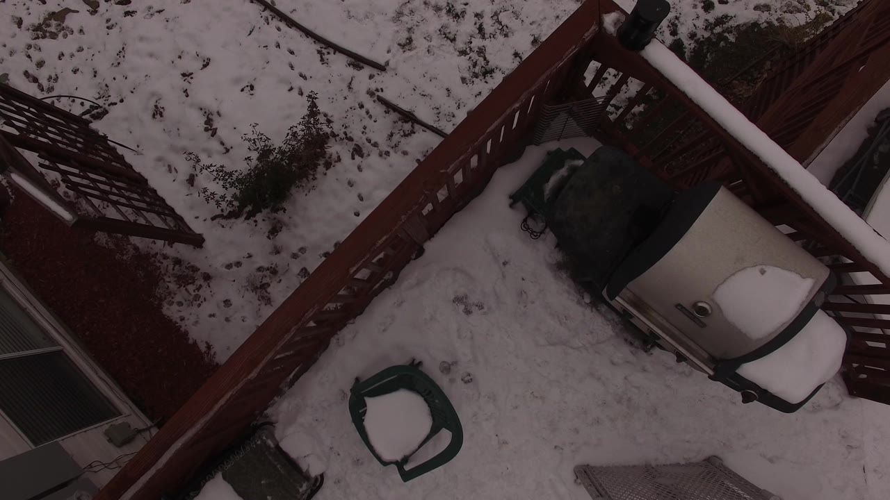 A slightly shifted overhead frame shows the Jefferson County patio, with the covered grill near the deck rail and a small green seat on the snow, no visible people.