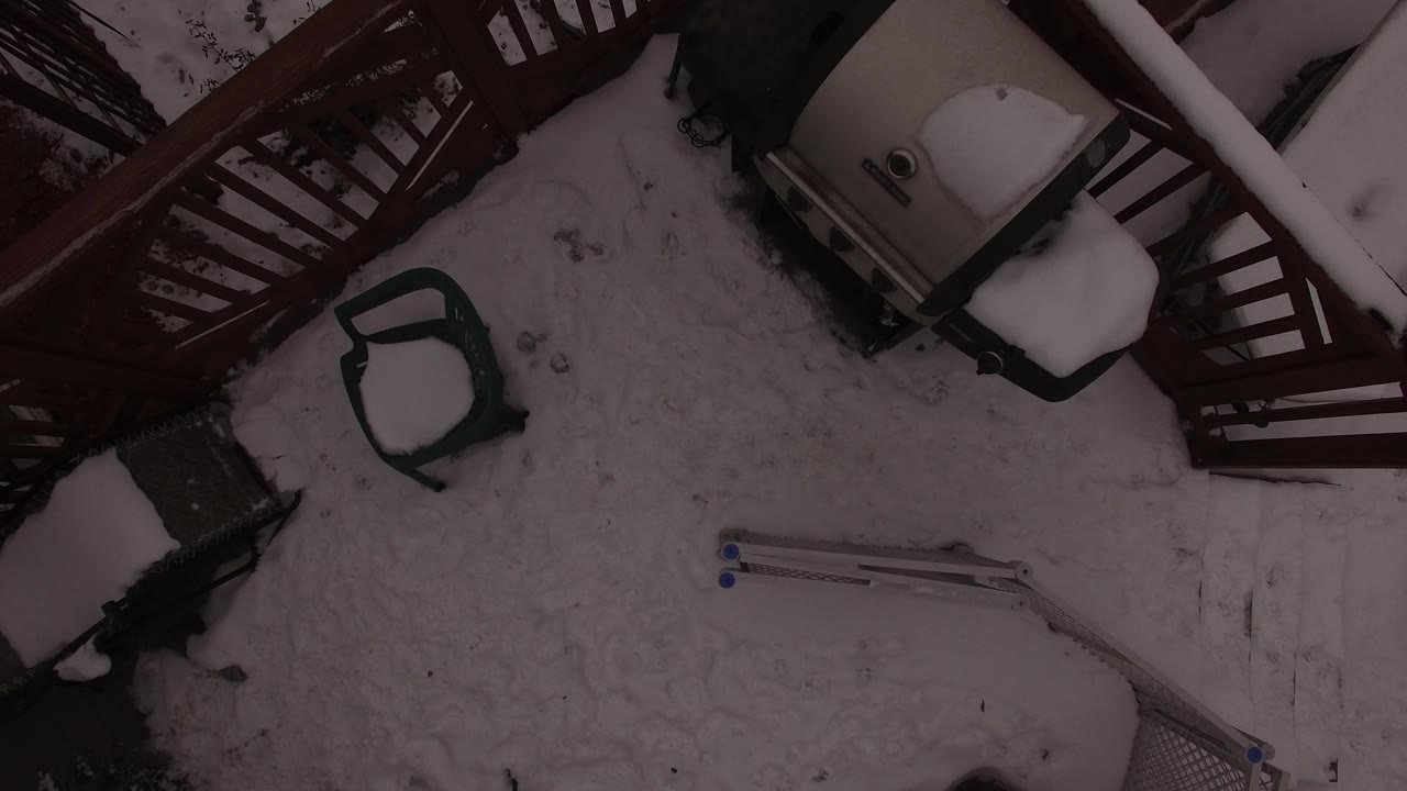 Overhead Jefferson County, Colorado backyard frame showing snow across the patio area, deck stairs at the top, a green chair near center left, and no visible people.