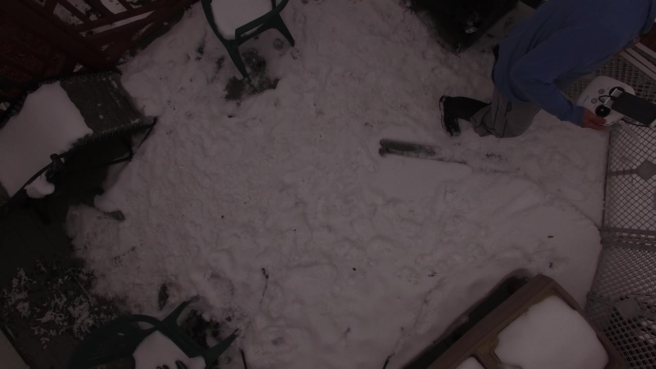 Dim overhead frame of a snowy backyard in Jefferson County, Colorado, with patio chairs, deck railing, a small snow-covered structure, and a partially visible man at the upper edge.