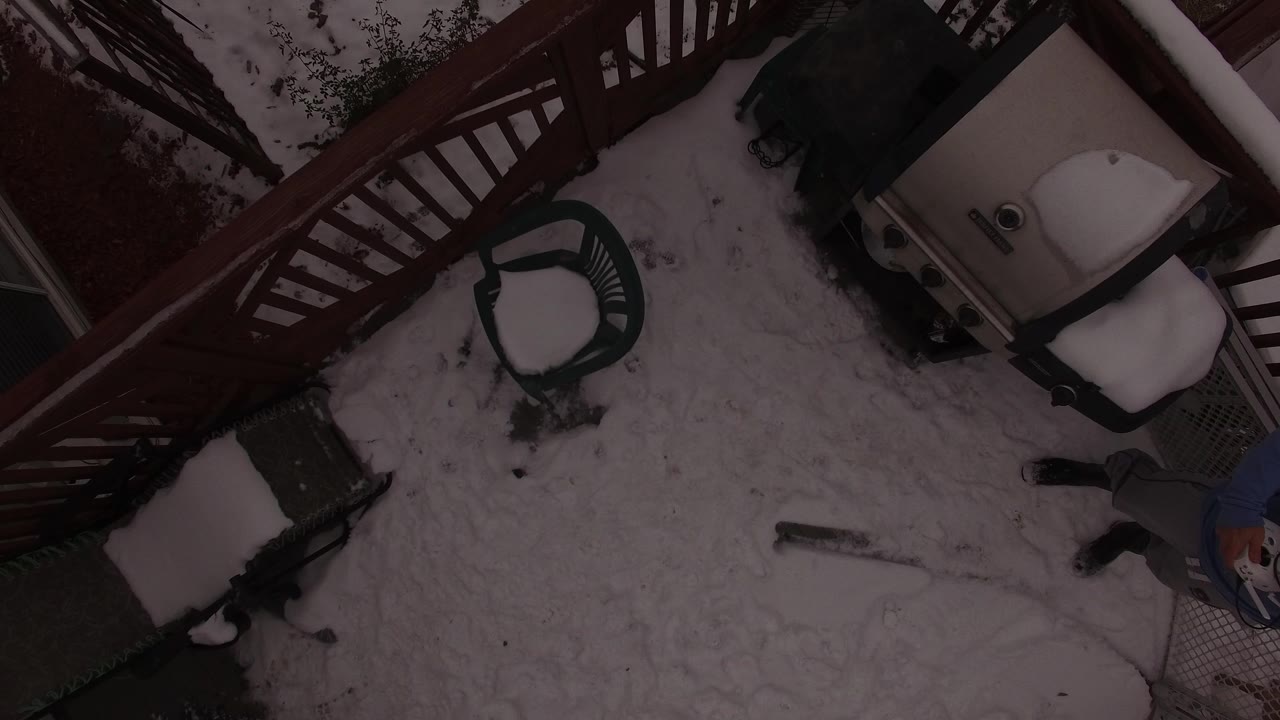Overhead frame in Jefferson, Colorado showing snow-covered patio furniture, red deck railing, a covered grill or patio appliance, and a person partly visible along the lower right edge.