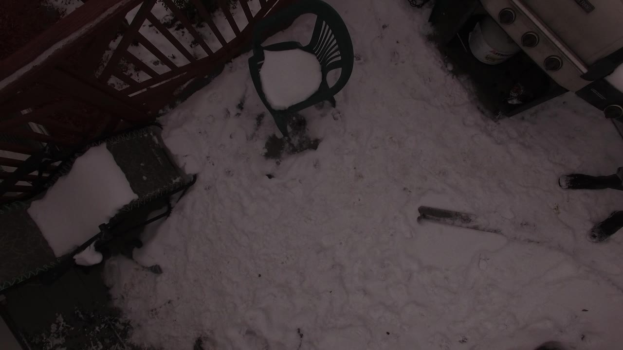 A tilted overhead drone view in Jefferson County, Colorado shows snow-covered ground beside a deck railing and outdoor chairs, with no visible people.