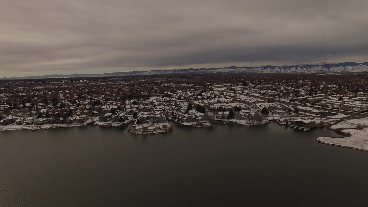 Wide aerial view over dark lake water toward snowy residential shoreline and distant hills.