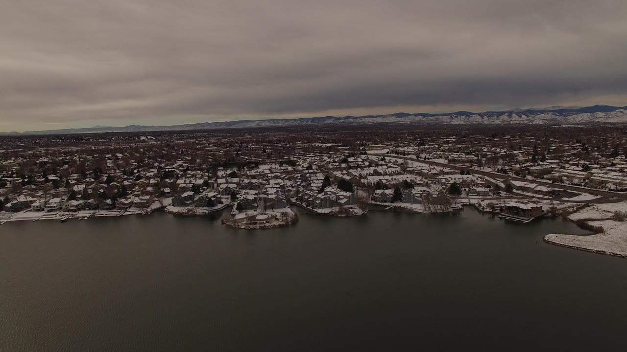 Aerial winter lake view in Jefferson County, Colorado, with dark water below, snow on the shoreline neighborhoods, low cloud cover, and no visible people.