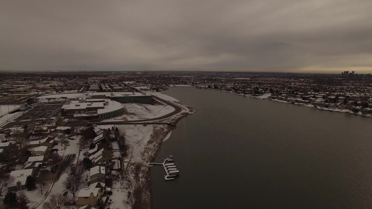 Aerial frame over Jefferson, Colorado with a curved snowy shoreline, residential roofs in the foreground, open water across the middle, and no visible people.