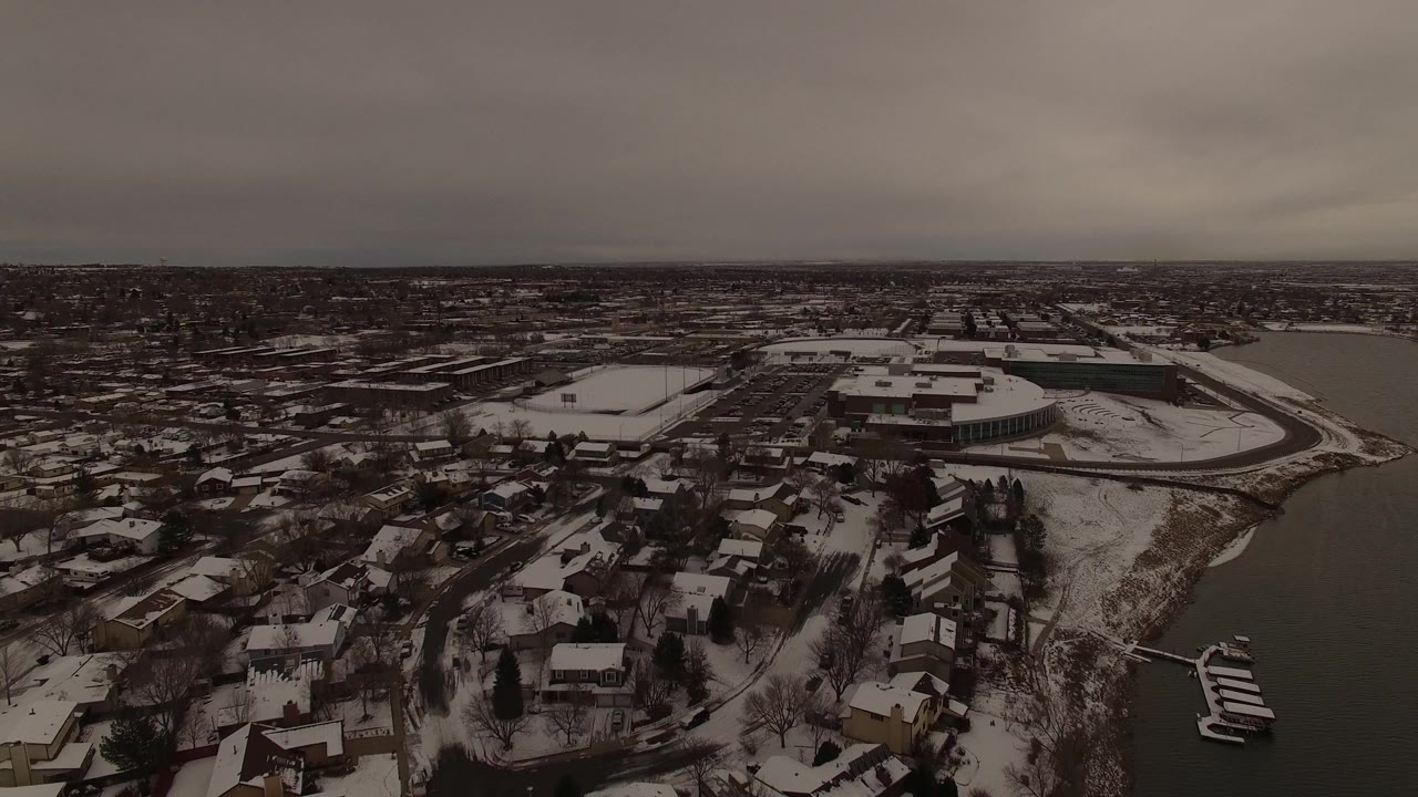 Aerial frame in Jefferson County, Colorado showing a dense snowy neighborhood, a curved water edge to the right, low institutional-looking buildings beyond it, and no visible people.