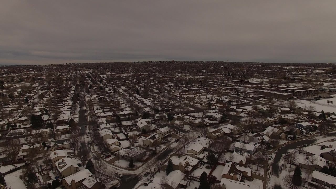 Wide aerial frame over Jefferson County, Colorado homes with snow on rooftops, dark streets, leafless trees, and no visible people.