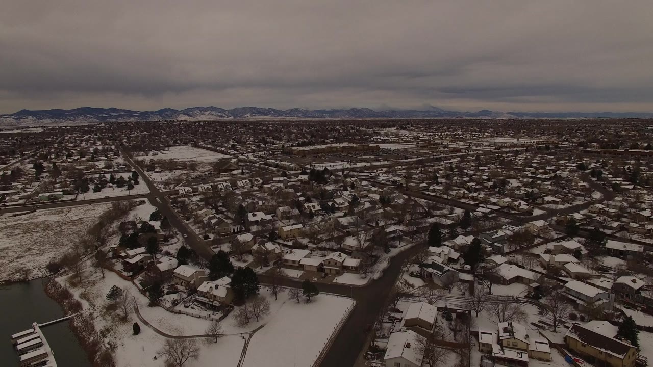 High view across Jefferson County, Colorado homes with snow on roofs and yards, a curved lake shoreline in the foreground, distant hills, and no visible people.