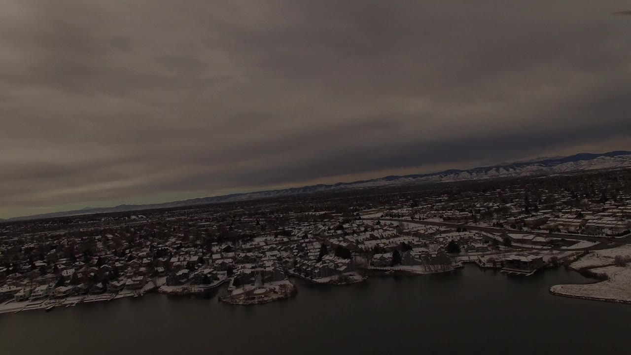 Aerial frame over a dark lake toward snow-dusted homes and trees in Jefferson County, Colorado, with distant low mountains and no visible people.