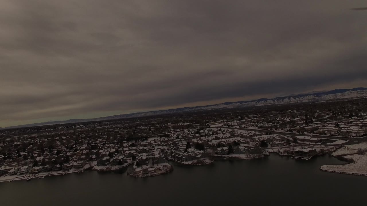 Wide aerial view of a winter neighborhood beside a lake under gray clouds.