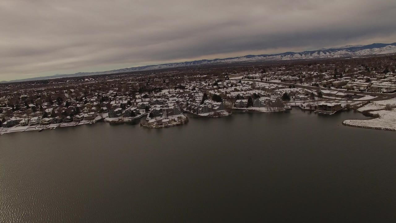 Wide aerial view over dark water toward a snowy residential shoreline under clouds.