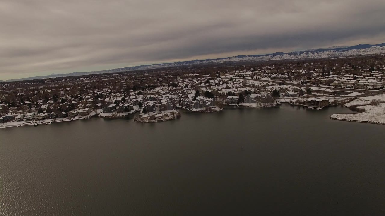 Aerial view over dark water toward snowy homes and distant mountains.