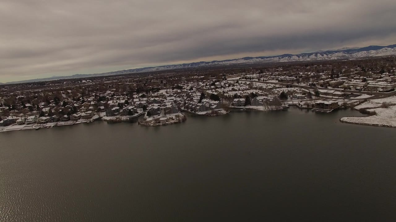 Aerial view in Jefferson County, Colorado, with rippled dark water in the foreground, clustered snowy homes along the shoreline, and mountains stretching across the cloudy background, with no visible people.
