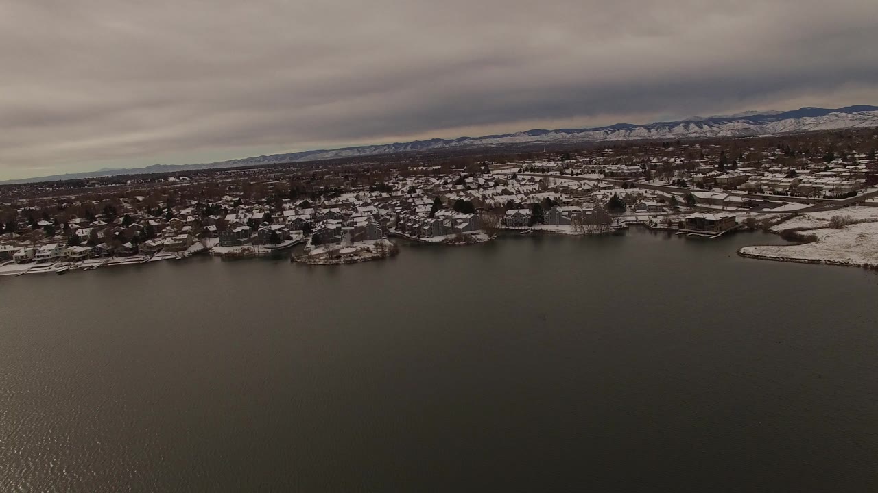 Aerial frame in Jefferson County, Colorado looking across dark lake water toward snow-lined homes, docks, distant mountains, and a gray overcast sky, with no visible people.