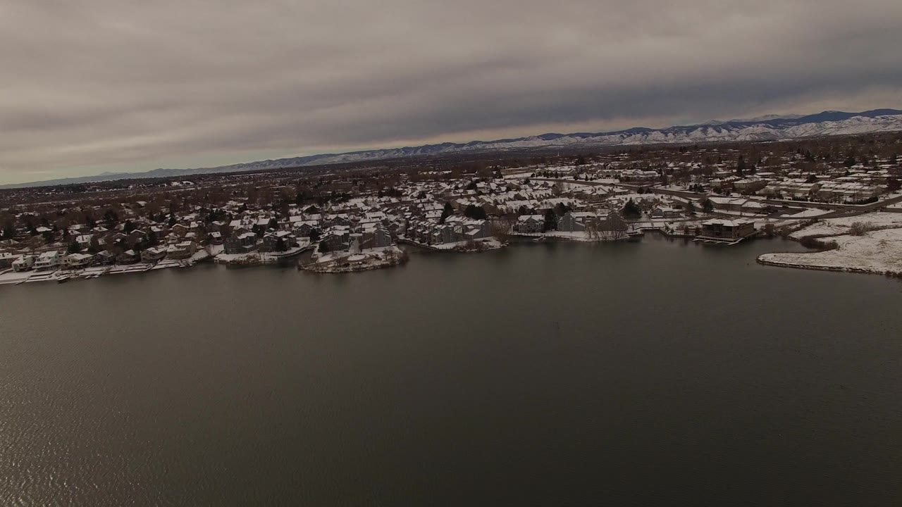 Wide aerial view in Jefferson County, Colorado with the lake occupying most of the foreground and a band of snowy homes and docks below distant mountains, with no visible people.