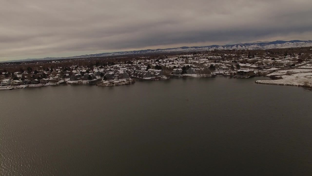 Opening aerial frame over gray lake water in Jefferson County, Colorado, with snowy shoreline homes, distant mountains, heavy clouds, and no visible people.