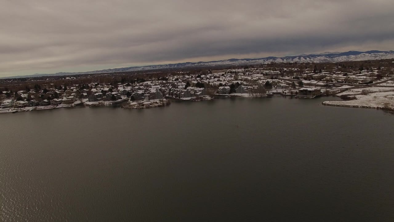 Wide aerial view in Jefferson County, Colorado, showing dark gray lake water, a snow-dusted residential shoreline, mountains on the horizon, and no visible people.