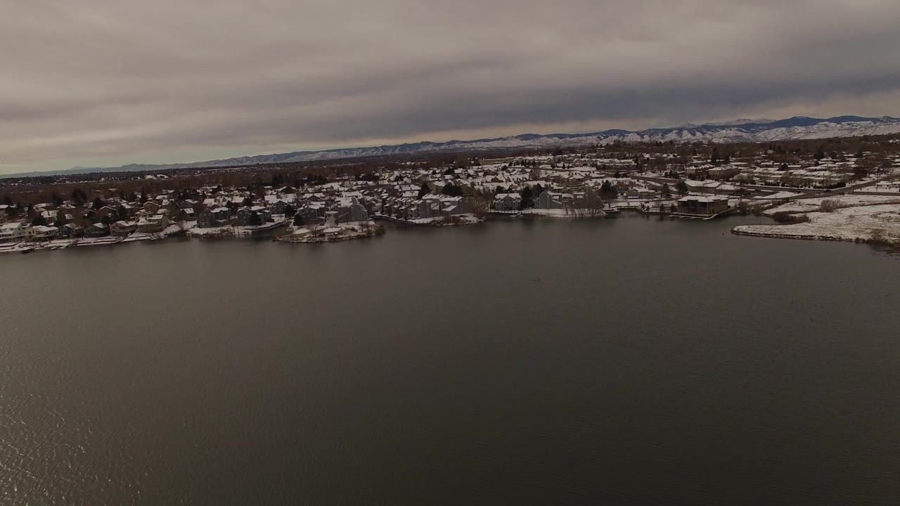 Snowy shoreline community seen beyond broad lake water under gray clouds.