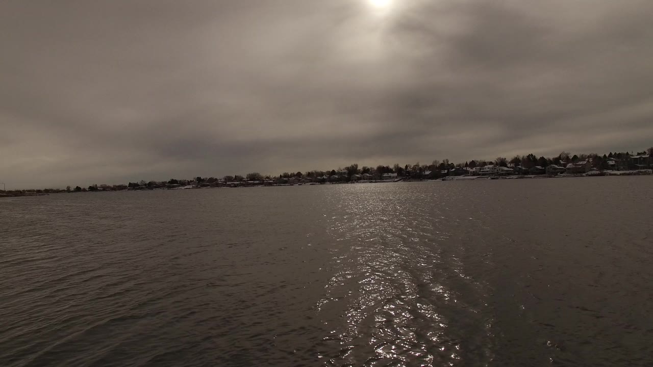 Wide Jefferson, Colorado water view with the bright reflection shifting near the lower center, heavy clouds above, distant shoreline homes, and no visible people.
