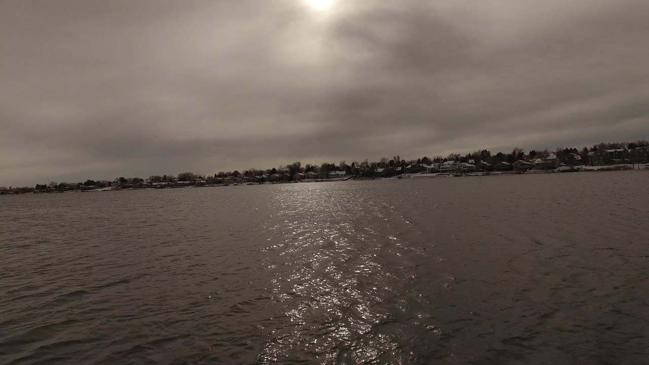 At 0:01, the water surface remains dominant as sun glare stretches toward the camera, with Jefferson, Colorado shoreline homes and trees visible under gray clouds and no visible people.