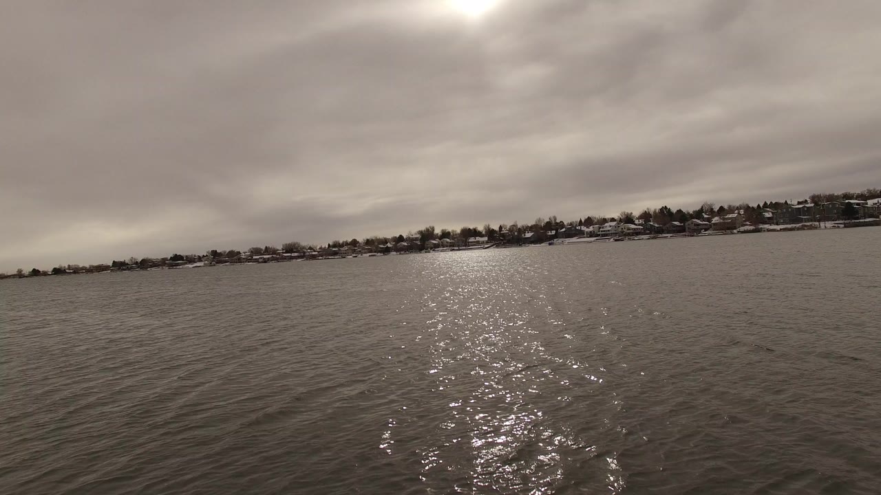 Sun glare on rippled lake water with distant snowy shoreline houses.