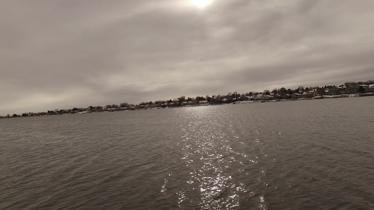 Jefferson, Colorado lake surface with the sun reflection centered more strongly across the ripples, distant snow-lined homes on the horizon, muted clouds, and no visible people.