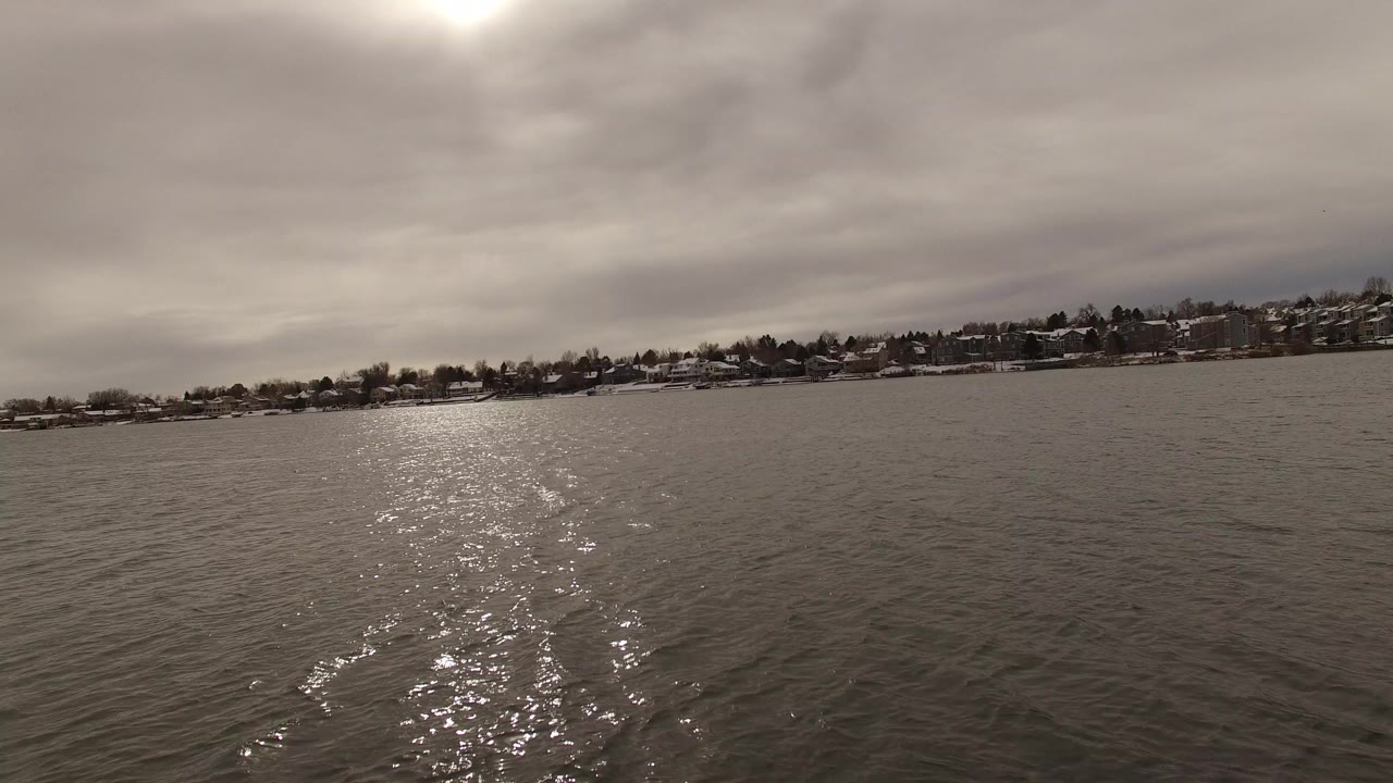 Opening view in Jefferson, Colorado showing rippled water, a bright glare path below the clouded sun, distant snow-covered homes, and no visible people.