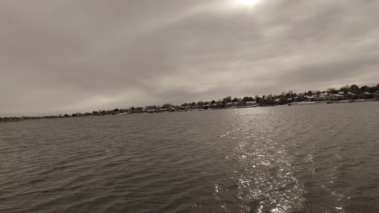 Open water fills the frame in Jefferson, Colorado, with distant houses and trees along the shoreline, a low horizon, strong reflected light, and no visible people.
