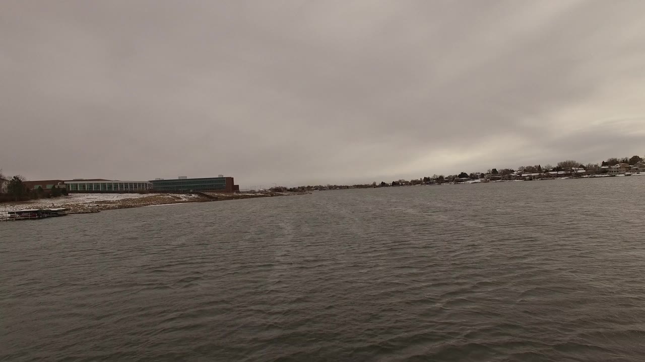 Wide lake water with snowy shoreline and low buildings under gray clouds.